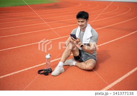 Asian athletic man using smartphone while resting after running on racetrack in sports stadium Asian athletic man using smartphone while resting after running on racetrack in sports stadium 141377138