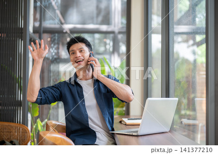 Asian entrepreneur man waving a hand while talking to client on smartphone during business call 141377210
