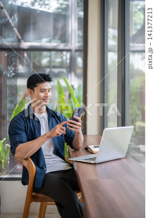 Asian man using smartphone while sitting with laptop at cafe table. Asian man using smartphone while sitting with laptop at cafe table. 141377213