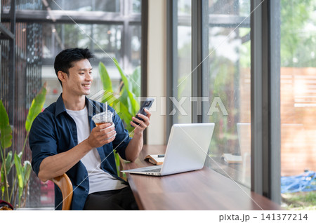 Asian freelancer man drinking iced coffee while using smartphone and laptop Asian freelancer man drinking iced coffee while using smartphone and laptop 141377214