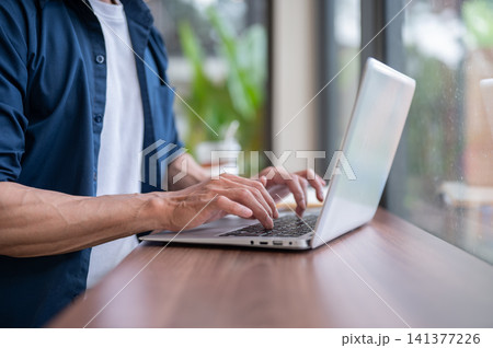 Close up of asian man's hands typing on laptop at wooden table while working in the cafe. Close up of asian man's hands typing on laptop at wooden table while working in the cafe. 141377226