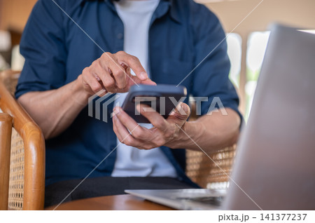 Close up of a freelancer adult man's hand using smartphone beside laptop while sitting at cafe table Close up of a freelancer adult man's hand using smartphone beside laptop while sitting at cafe table 141377237