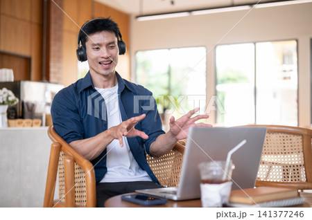 Asian man wearings headphones speaking happily during online meeting with laptop in modern cafe. Asian man wearings headphones speaking happily during online meeting with laptop in modern cafe. 141377246