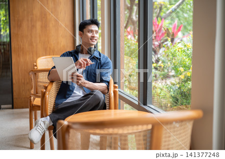 Asian man artist holding tablet and stylus pen looking over the glass wall while sitting in cafe. Asian man artist holding tablet and stylus pen looking over the glass wall while sitting in cafe. 141377248