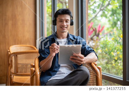Asian artist man using a tablet and enjoy listening to music in headphones while working in the cafe Asian artist man using a tablet and enjoy listening to music in headphones while working in the cafe 141377255