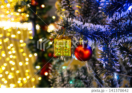 Close up red ball and golden present box hanging on pine tree branch with blurry warm yellow bokeh background for christmas celebration 141378642