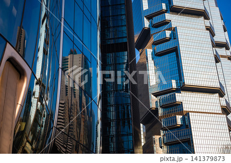 Modern blue geometric glass skyscraper architecture features complex symmetrical window facade reflecting bright sunlight standing alongside tall corporate business office tower structure Modern blue geometric glass skyscraper architecture features complex symmetrical window facade reflecting bright sunlight standing alongside tall corporate business office tower structure 141379873