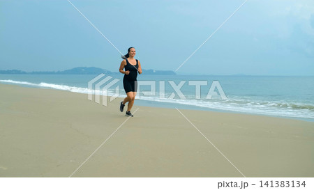Young woman in black athletic outfit running energetically along a beach smiling with sea and tropical hills visible behind. Energetic workout concept Young woman in black athletic outfit running energetically along a beach smiling with sea and tropical hills visible behind. Energetic workout concept 141383134