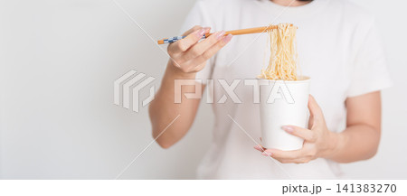 Happy young Asian woman enjoying a delicious cup of hot instant noodles while relaxing at home, Cheerful female eating instant ramen with a smile at apartment. Simple lifestyle and quick meal concept Happy young Asian woman enjoying a delicious cup of hot instant noodles while relaxing at home, Cheerful female eating instant ramen with a smile at apartment. Simple lifestyle and quick meal concept 141383270