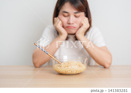 Bored young Asian woman eating instant noodles at home. Exhausted female looking unhappy while eating bowl ramen. quick meal after work, burnout and unhealthy eating habit, lifestyle and cheap meal Bored young Asian woman eating instant noodles at home. Exhausted female looking unhappy while eating bowl ramen. quick meal after work, burnout and unhealthy eating habit, lifestyle and cheap meal 141383281
