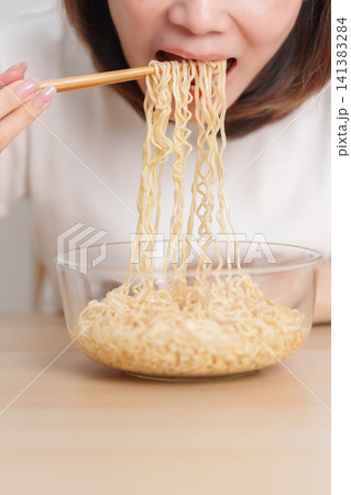Happy young Asian woman enjoying a delicious bowl of hot instant noodles while relaxing at home, Cheerful female eating instant ramen with a smile at apartment. Simple lifestyle and quick meal concept Happy young Asian woman enjoying a delicious bowl of hot instant noodles while relaxing at home, Cheerful female eating instant ramen with a smile at apartment. Simple lifestyle and quick meal concept 141383284