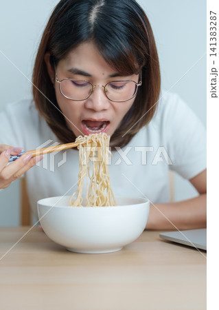 Happy young Asian woman working late at night on laptop while enjoying a bowl of hot instant ramen noodles at home. Cheerful female having a quick meal during work from home. Simple lifestyle concept 141383287