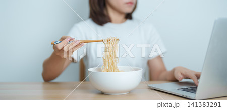 Happy young Asian woman working late at night on laptop while enjoying a bowl of hot instant ramen noodles at home. Cheerful female having a quick meal during work from home. Simple lifestyle concept Happy young Asian woman working late at night on laptop while enjoying a bowl of hot instant ramen noodles at home. Cheerful female having a quick meal during work from home. Simple lifestyle concept 141383291