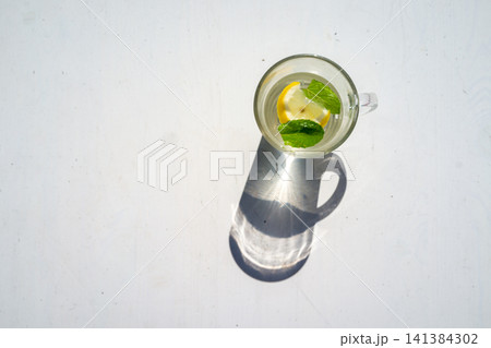 Top view of clear citrus drink in glass mug with lemon slice and mint leaves, casting long shadow on bright white surface, minimalist refreshment with clean summer feeling Top view of clear citrus drink in glass mug with lemon slice and mint leaves, casting long shadow on bright white surface, minimalist refreshment with clean summer feeling 141384302