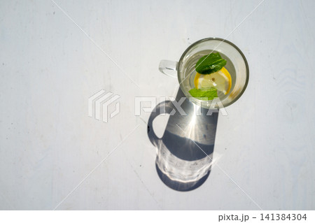 Top view glass mug with lemon slice and mint leaves in clear water on bright white background, casting strong shadow and sunlight reflection, minimal refreshing drink composition Top view glass mug with lemon slice and mint leaves in clear water on bright white background, casting strong shadow and sunlight reflection, minimal refreshing drink composition 141384304