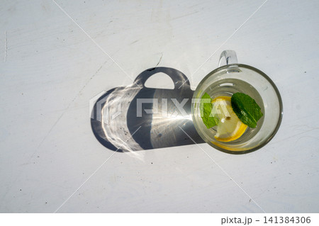 Top view of glass mug with lemon slice and fresh mint leaves casting strong sunlit shadow on white surface, minimal refreshing drink composition with bright natural light and clean seasonal wellness Top view of glass mug with lemon slice and fresh mint leaves casting strong sunlit shadow on white surface, minimal refreshing drink composition with bright natural light and clean seasonal wellness 141384306