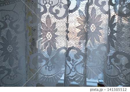 White lace curtains in window. Bright light through intricate floral design creates soft glow 141385512