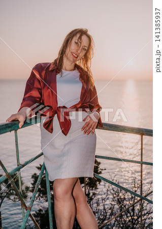 Woman sunset ocean happy middle aged woman standing on a balcony overlooking the sea at sunset during a summer vacation trip 141385937