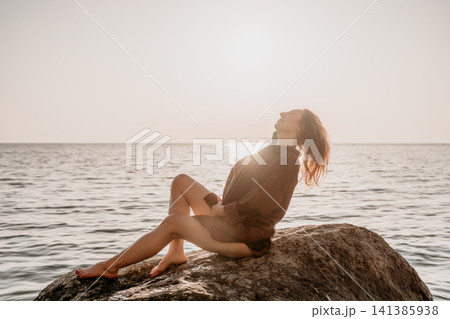 Woman beach sunset young woman sitting on a rock by the sea at sunset enjoying the golden hour Woman beach sunset young woman sitting on a rock by the sea at sunset enjoying the golden hour 141385938