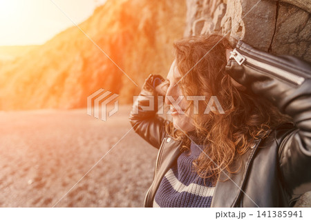 Woman leather jacket sunset relaxed young woman leaning against a stone wall at the beach during golden hour Woman leather jacket sunset relaxed young woman leaning against a stone wall at the beach during golden hour 141385941