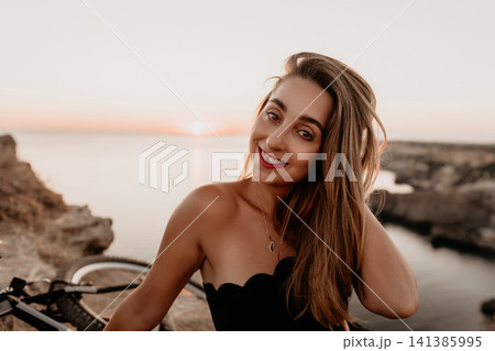 Woman sunset coast smiling young lady sitting on cliffs by the ocean during golden hour with a bicycle Woman sunset coast smiling young lady sitting on cliffs by the ocean during golden hour with a bicycle 141385995