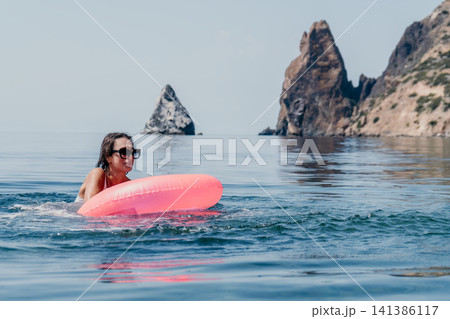 Swimming woman sea with pink inflatable ring enjoying summer vacation near dramatic coastal rocks Swimming woman sea with pink inflatable ring enjoying summer vacation near dramatic coastal rocks 141386117