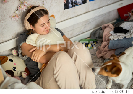 Teen girl in messy bedroom with dog listening to music Teen girl in messy bedroom with dog listening to music 141389139