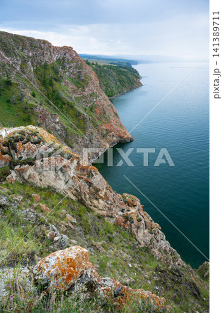 Vertical landscape of Lake Baikal, a steep rocky cliff covered in orange lichen dropping into deep blue water. Rugged coastline of Olkhon Island in summer Vertical landscape of Lake Baikal, a steep rocky cliff covered in orange lichen dropping into deep blue water. Rugged coastline of Olkhon Island in summer 141389711