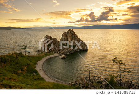 Wide landscape view of a jagged Shamanka rock in a still Lake Baikal at sunset. Golden hour sunlight reflecting on water with a curved beach, Russia Wide landscape view of a jagged Shamanka rock in a still Lake Baikal at sunset. Golden hour sunlight reflecting on water with a curved beach, Russia 141389713