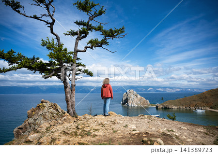 Woman standing on a cliff looking at Lake Baikal. Rear view of a female tourist in a red hoodie near Shamanka Rock on Olkhon Island. Outdoor travel and adventure concept Woman standing on a cliff looking at Lake Baikal. Rear view of a female tourist in a red hoodie near Shamanka Rock on Olkhon Island. Outdoor travel and adventure concept 141389723