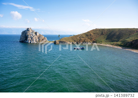 Summer landscape of Lake Baikal with Shamanka Rock and a blue boat in the bay. Grassy peninsula and clear sky during daytime, Olkhon Island in Russia Summer landscape of Lake Baikal with Shamanka Rock and a blue boat in the bay. Grassy peninsula and clear sky during daytime, Olkhon Island in Russia 141389732