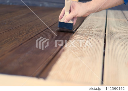 Brown oil for protection of wood by staining. Painting a larch for protecting and pests. Woodworker applying finish to terrace deck board. Repairman varnishing with brush wooden plank floor surface. Brown oil for protection of wood by staining. Painting a larch for protecting and pests. Woodworker applying finish to terrace deck board. Repairman varnishing with brush wooden plank floor surface. 141390162