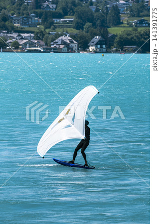 Person wing foiling on a lake with a town in the background, illustrating eco friendly wind driven recreation and water sports. Person wing foiling on a lake with a town in the background, illustrating eco friendly wind driven recreation and water sports. 141391775