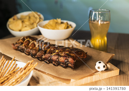 Barbecue ribs on a wooden board, beer and snacks arranged for a soccer viewing party, creating a cozy football night atmosphere. Barbecue ribs on a wooden board, beer and snacks arranged for a soccer viewing party, creating a cozy football night atmosphere. 141391780