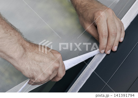 Person adjusting a mosquito screen on hook and loop tape on a window frame, illustrating home maintenance and practical DIY work. Person adjusting a mosquito screen on hook and loop tape on a window frame, illustrating home maintenance and practical DIY work. 141391794