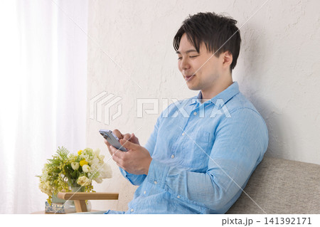 スマホを見ながらソファでくつろぐ若い男性 スマホを見ながらソファでくつろぐ若い男性 141392171
