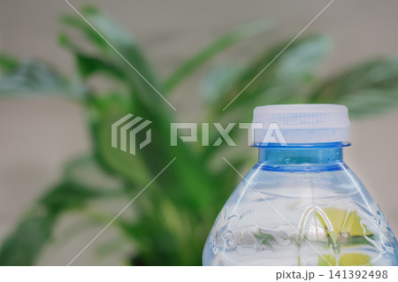 Close up view of transparent blue plastic water bottle with white tethered cap compliant with European Union eco regulations on blurred green plant background 141392498