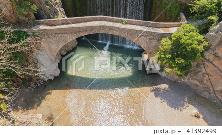 High aerial view of the historic stone arch bridge over a clear river stream with Paleokaria waterfall in Thessaly Greece on a bright sunny summer day High aerial view of the historic stone arch bridge over a clear river stream with Paleokaria waterfall in Thessaly Greece on a bright sunny summer day 141392499