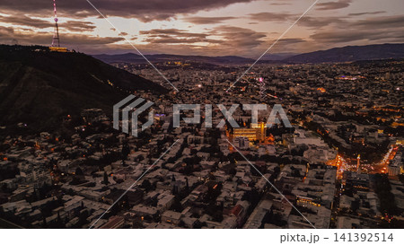 Aerial view of Tbilisi cityscape at dusk featuring Mtatsminda mountain with illuminated TV tower under a cloudy purple sunset sky in Georgia South Caucasus Aerial view of Tbilisi cityscape at dusk featuring Mtatsminda mountain with illuminated TV tower under a cloudy purple sunset sky in Georgia South Caucasus 141392514