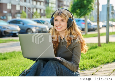 Smiling young woman in headphones sitting on grass and using laptop city 141392573