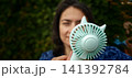 Portable pocket fan held by a person in a green outdoor area on a sunny day 141392784