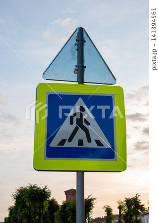Pedestrian crossing sign with triangular warning sign above. Yellow border frames blue symbol of walking person. Sign mounted on metal pole outdoors. Sky shows soft clouds at dusk or dawn Pedestrian crossing sign with triangular warning sign above. Yellow border frames blue symbol of walking person. Sign mounted on metal pole outdoors. Sky shows soft clouds at dusk or dawn 141394561
