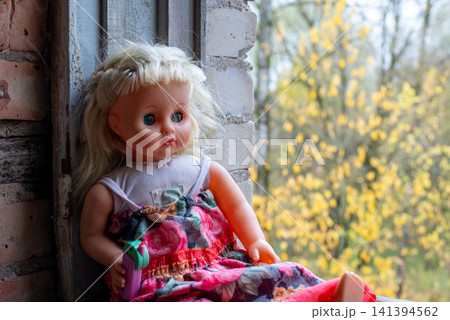 Blonde doll leans against brick window frame. Soft-focus yellow trees frame the background. Pink floral dress with green accents. Doll wears simple white top underneath 141394562