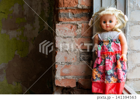 Doll stands beside weathered brick wall. Childlike figure wears floral dress. Dress has red hem and colorful pattern. Background shows peeling green paint. Rustic wall suggests aged, abandoned setting Doll stands beside weathered brick wall. Childlike figure wears floral dress. Dress has red hem and colorful pattern. Background shows peeling green paint. Rustic wall suggests aged, abandoned setting 141394563