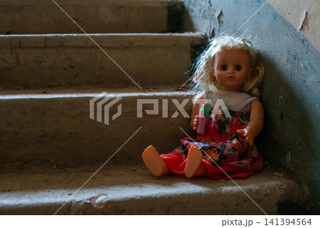 Doll sits alone on dusty concrete stairs. Blonde hair and colorful dress are visible. Soft lighting highlights worn, aged appearance. Staircase shows signs of neglect and time. Small figure adds eerie Doll sits alone on dusty concrete stairs. Blonde hair and colorful dress are visible. Soft lighting highlights worn, aged appearance. Staircase shows signs of neglect and time. Small figure adds eerie 141394564