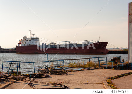 Large red cargo ship rests at harbor dockside. Sunlight casts warm glow over water and vessel. Rusted metal railings frame foreground view. Docks show signs of weathering and neglect 141394579