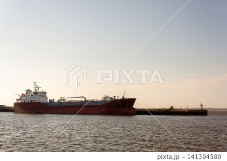 Large red cargo ship floats calmly on water. Sunset hues glow softly behind vessel. Ship moored near dark pier structure. Clear sky and calm sea frame the scene Large red cargo ship floats calmly on water. Sunset hues glow softly behind vessel. Ship moored near dark pier structure. Clear sky and calm sea frame the scene 141394580
