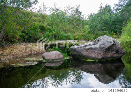 Still water reflects mossy rocks beside green forest. Vegetation grows thick on grassy bank. Large boulders rest at water's edge. Foliage and sky mirror in calm surface. Natural landscape shows quiet Still water reflects mossy rocks beside green forest. Vegetation grows thick on grassy bank. Large boulders rest at water's edge. Foliage and sky mirror in calm surface. Natural landscape shows quiet 141394622
