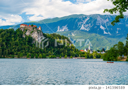 Panoramic view of Lake Bled in Slovenia with Bled Castle overlooking the lake, surrounded by forests and the majestic Julian Alps under a clear blue sky. 141396589