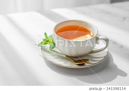 Cup of tea with lemon and mint on a marble background with spoon near window Cup of tea with lemon and mint on a marble background with spoon near window 141398114